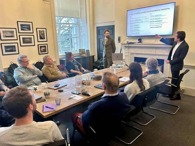 A group seating around a meeting table listening to two presenters pointing at their slides on the board
